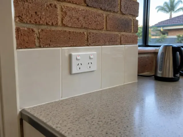New double power point fitted into a tiled kitchen splashback beside a brick wall and window in Bundall